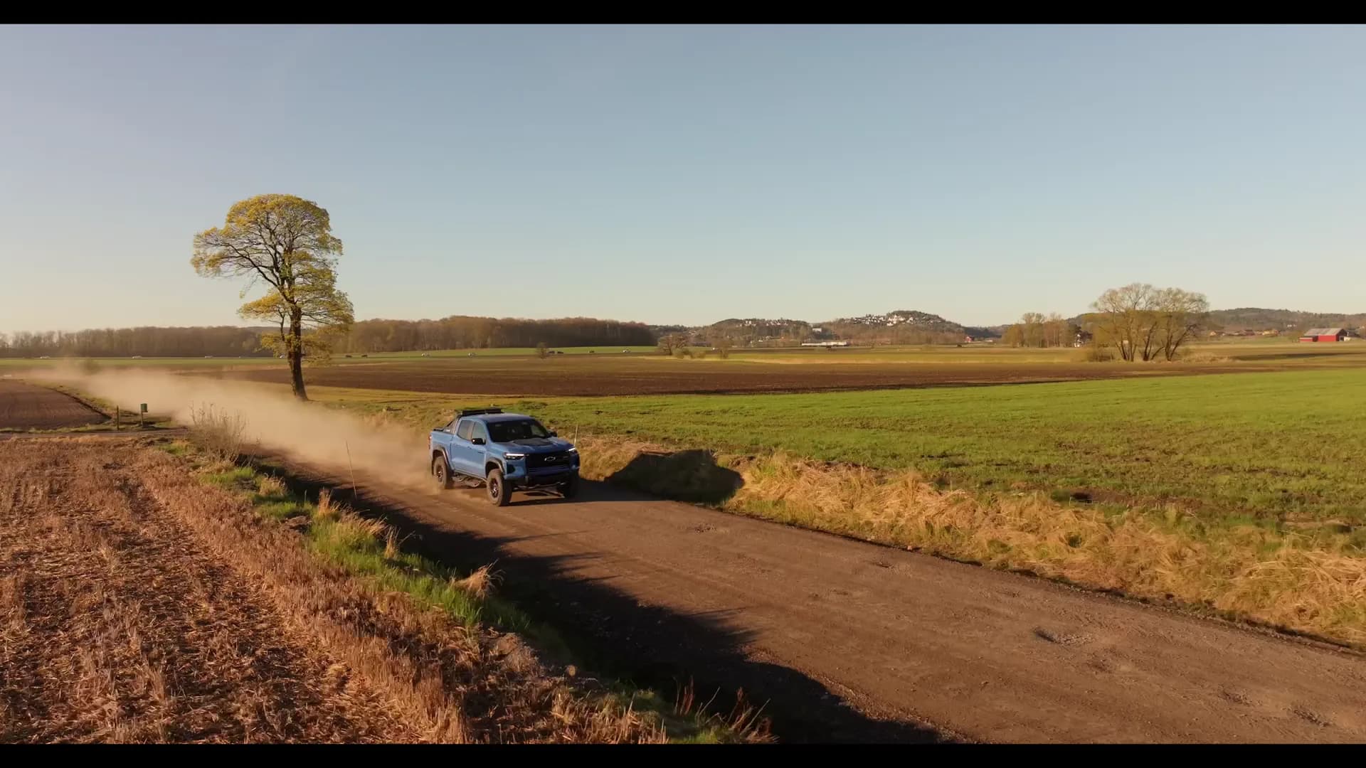 En blå Chevrolet Colorado ZR2 på en grusvei mellom jordene, med støv bak seg og et ensomt tre i bakgrunnen.