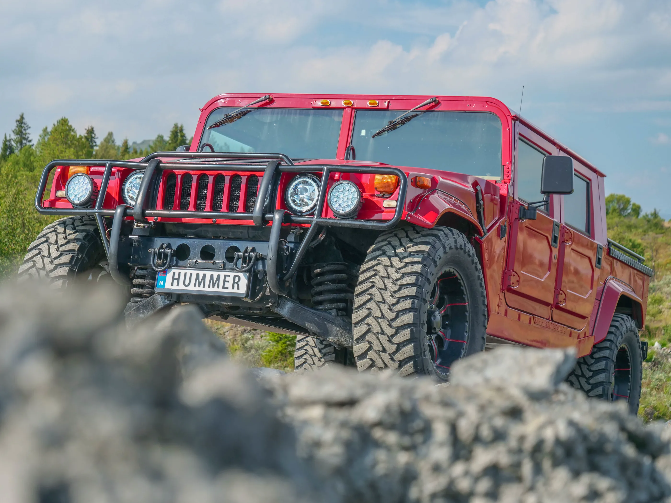 Rød Hummer H1 på en grusrygg med skog i bakgrunnen.