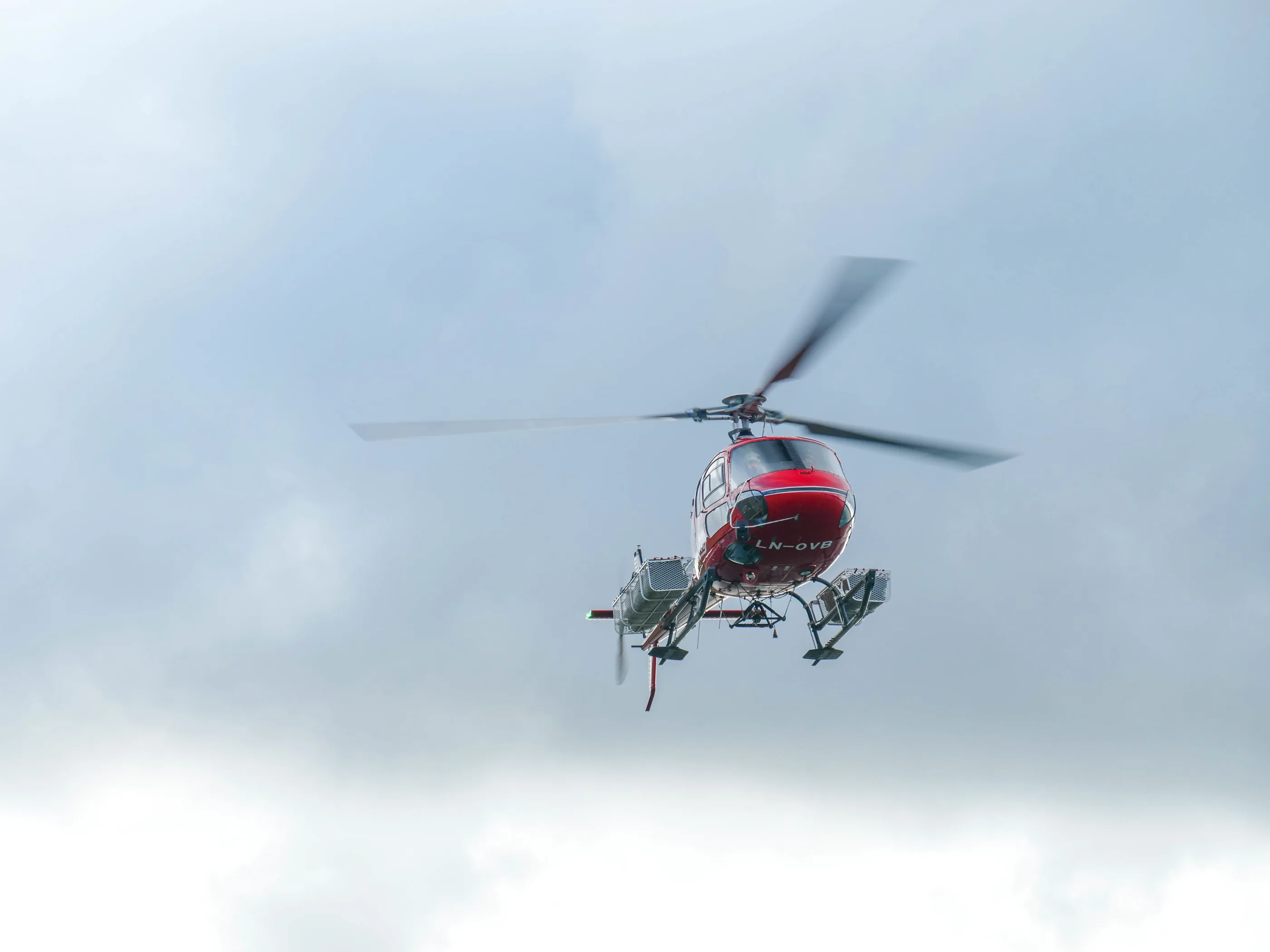 Rødt helikopter i lufta mot en grå himmel.