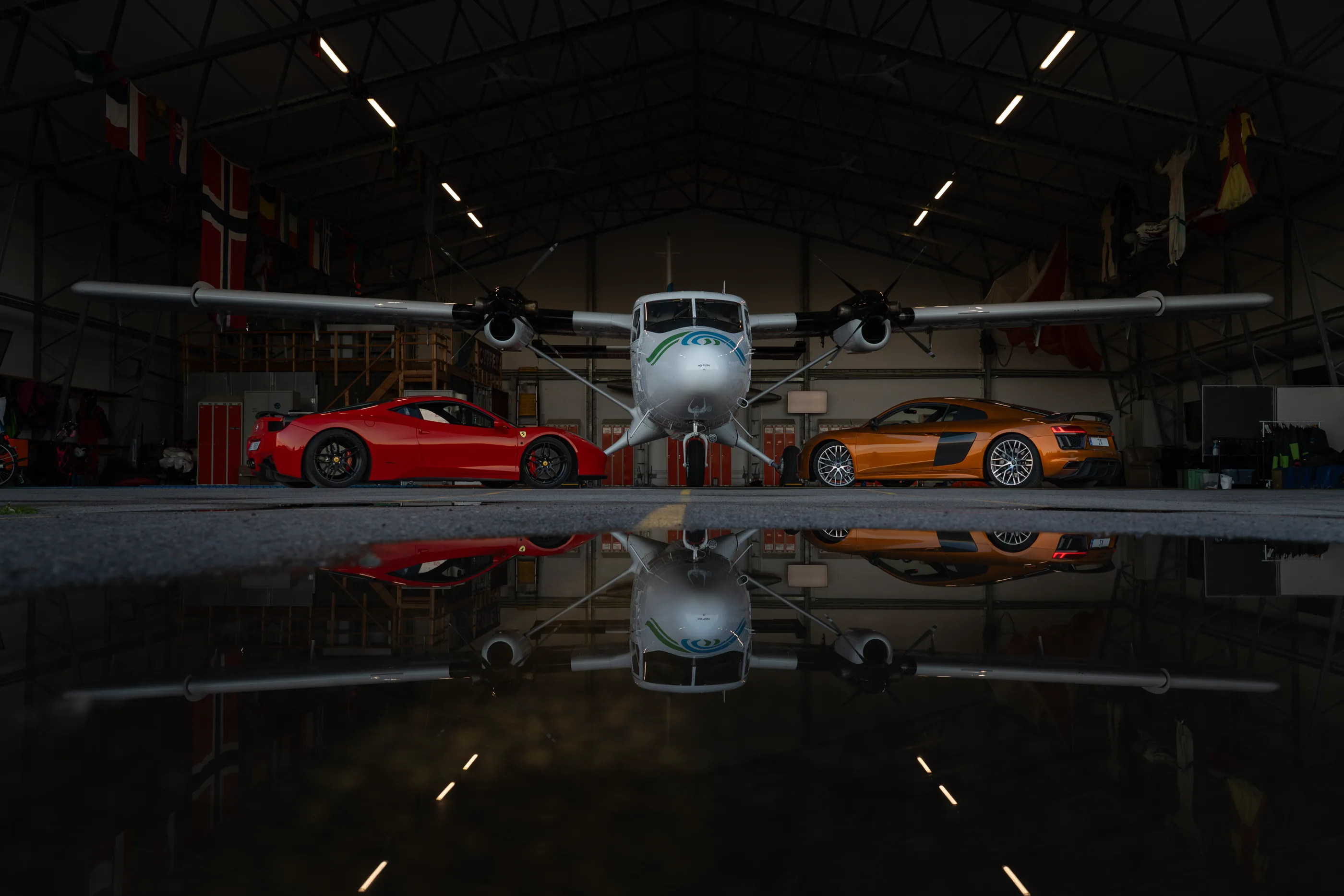 Fly og to sportsbiler i en hangar, speilet i det våte gulvet.
